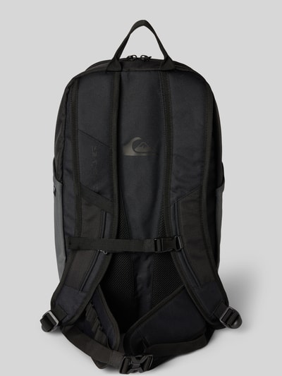 Männermodel trägt Rucksack mit Logo-Patch Modell 'ALTACAMA' von Quiksilver in Schwarz mit verstellbaren Schultergurten und Tragegriff, Ganzkörperansicht Rückansicht.