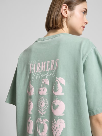 Frauenmodel trägt Oversized Damen T-Shirt von OH APRIL aus reiner Baumwolle, Modell 'Farmers Market' mit Label-Stitching, Rückenlänge 69 cm, mintgrün mit rosa Print, Ganzkörperansicht Rückansicht.