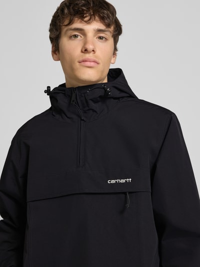 Männermodel trägt schwarzen Windbreaker mit Label-Stitching von Carhartt Work In Progress, Regular-Fit, Kapuze, Reißverschluss, Nahaufnahme Oberkörper, Rückenlänge bei Größe M 73 cm, Schulterbreite 48 cm, Brustweite 55 cm, Ärmellänge 66 cm.