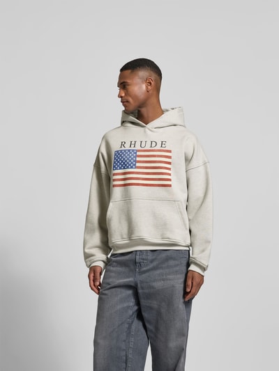 Männermodel trägt Oversized Hoodie mit Logo-Print von RHUDE in Beige mit US-Flagge, reine Baumwolle, Känguru-Tasche, Ganzkörperansicht von vorne.