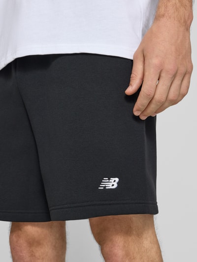 Mężczyzna nosi Relaxed Fit Shorts mit Logo-Stitching od New Balance, mieszanka bawełny, czarne, wyhaftowane logo, szerokość w rozmiarze M 42 cm, długość wewnętrzna nogawki w rozmiarze M 18 cm, detal, zbliżenie na dolną część ciała.