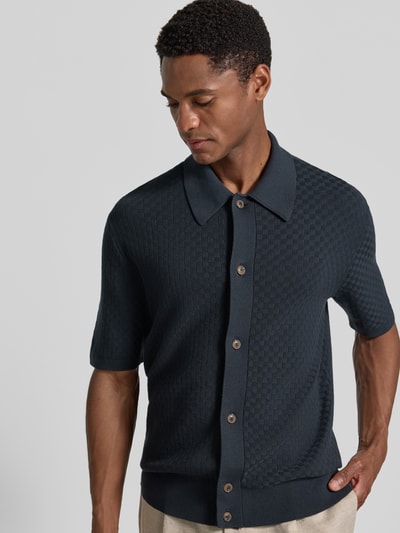 Męski model nosi koszulkę polo Regular Fit Poloshirt mit Strukturmuster Modell 'ARAAWEN' von Tiger Of Sweden z bawełny, ciemny kolor, kołnierzyk polo, fakturowany wzór, krótki rękaw, listwa guzikowa, widok na górną część ciała z przodu.
