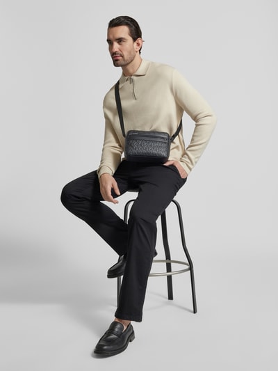 Männermodel trägt Handtasche mit Logo-Muster von Armani Exchange in Schwarz mit Label-Detail und Schulterriemen, Ganzkörperansicht von vorne.