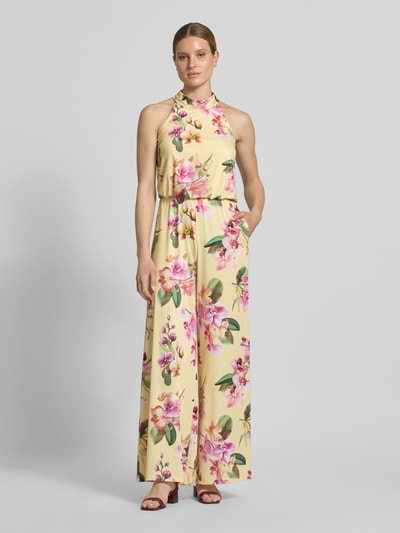Modelka nosi Regular Fit Jumpsuit mit floralem Allover-Print von V by Vera Mont w pastelowych kolorach, bez rękawów, krój regular fit, floralny wzór allover, cut-out na plecach, zamek wszyty w szwie, długość pleców w rozmiarze 36: 147 cm, szerokość w rozmiarze 36: 36 cm, całkowita długość w rozmiarze 36: 132 cm, długość nogawki wewnętrznej w rozmiarze 36: 81 cm, widok całego ciała z przodu.