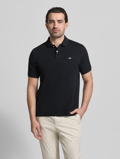 Męski model nosi Slim Fit Poloshirt mit Logo-Stitching od Gant, czarna bawełna, kołnierzyk polo, krótki rękaw, krój slim fit, widok z przodu, ujęcie całej sylwetki.