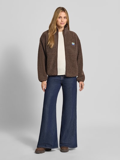 Frauenmodel trägt Regular Fit Jacke aus Teddyfell Modell 'Hoktown' von American Vintage in Braun mit Stehkragen und Logo-Patch, Ganzkörperansicht von vorne. Rückenlänge 62 cm, Schulterbreite 54 cm, Brustweite 46 cm, Ärmellänge 55 cm.