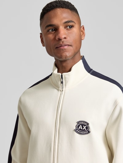 Mężczyzna nosi Sweatjacke mit Zweiwege-Reißverschluss Modell 'FELPA' od ARMANI EXCHANGE, bawełna z domieszką stretchu, krój regular fit, label-stitching, stójka, kolor kremowy z czarnymi wstawkami, zbliżenie na górę ciała, przód.