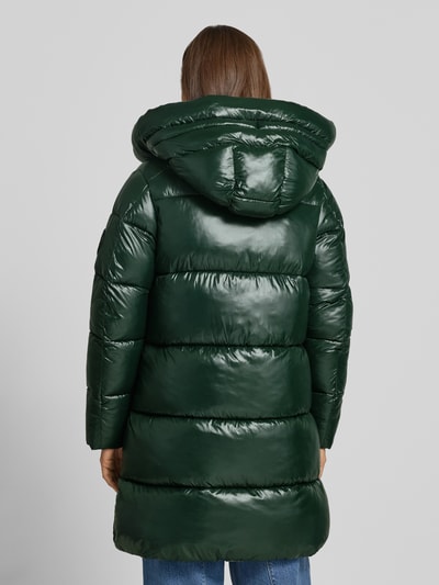 Frauenmodel trägt Steppmantel mit Kapuze und Label-Patch Modell 'ISABEL' von Save The Duck, glänzend grün, Ganzkörperansicht Rückansicht. Gerade geschnitten mit Tunnelzugkapuze, Zweiwege-Reißverschluss und seitlichen Reißverschlusstaschen.