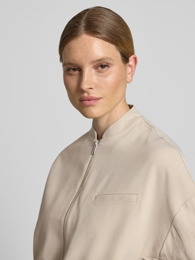 Modelka kobieca ma na sobie Blouson mit Brustleistentasche Modell 'Jamassi' od OPUS w kolorze beżowym, mieszanka materiałów z dodatkiem wiskozy, krój loose fit, stójka, zamek, kieszeń na piersi, tekstura gładka, zbliżenie górnej części ciała. Szerokość w rozmiarze 36: 62 cm, długość pleców: 53 cm, szerokość ramion: 59 cm, długość rękawa: 44 cm.