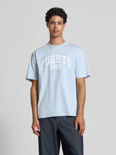 Männermodel trägt Relaxed Fit T-Shirt aus reiner Baumwolle Modell 'RICO' von Only & Sons in Hellblau mit weißem Motiv-Print, unifarben, Ganzkörperansicht von vorne.