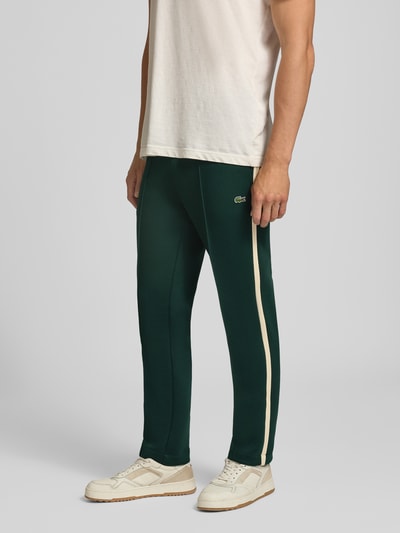 Männermodel trägt dunkelgrüne Regular Fit Jogpants aus Baumwoll-Mix von Lacoste, cremefarbene Seitenstreifen, Ganzkörperansicht von vorne. Herren Weite Hose von Lacoste.