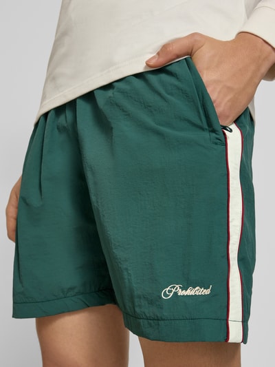 Männermodel trägt Shorts mit Label-Stitching Modell 'Slipstream' von PROHIBITED in Grün mit weiß-rotem Streifen, Nahaufnahme Oberschenkel, Regular Fit, elastischer Bund, Innenbeinlänge 16 cm, Bundweite 34 cm.