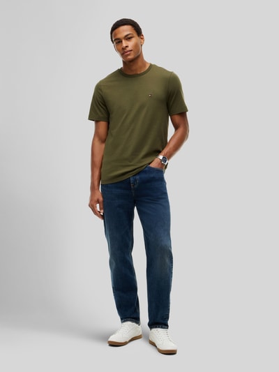 Męski model nosi Regular Fit T-Shirt aus reiner Baumwolle von Tommy Hilfiger w kolorze oliwkowym, gładka tekstura, pełny widok z przodu.