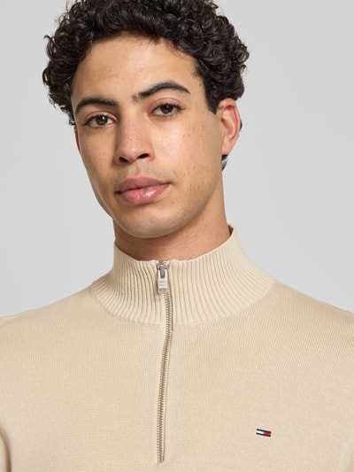 Männermodel trägt Slim Fit Troyer aus reiner Baumwolle von Tommy Jeans in Beige, Feinstrick, Logo-Detail, Nahaufnahme Oberkörper.