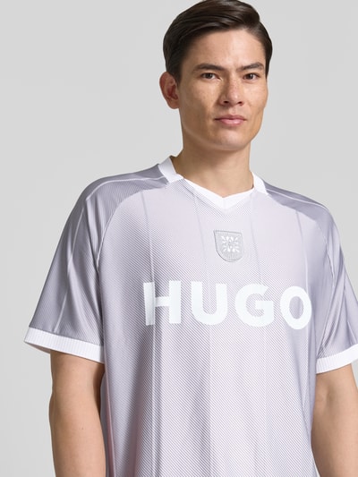 Männermodel trägt Regular Fit T-Shirt mit Label Print Modell 'DWILD' von HUGO, weiß-grau gestreift, V-Ausschnitt, Nahaufnahme Oberkörper.