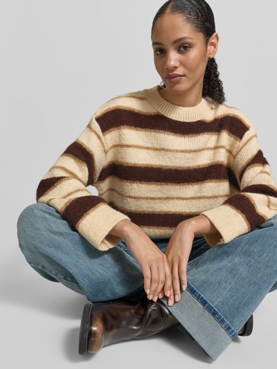 Frauenmodel trägt Strickpullover mit gerippten Abschlüssen von Mango im Material-Mix mit Baumwoll-Anteil, lockerer Schnitt, Rundhalsausschnitt, cremefarben mit braunen Streifen, jeans und braune Schuhe kombiniert, Ganzkörperansicht von vorne.