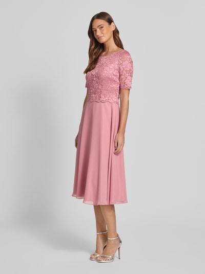 Modelka kobieca ma na sobie Knielanges Cocktailkleid mit Spitze und Rundhalsausschnitt od Vera Mont z szyfonu, linia A, długość do kolan, koronka, okrągły dekolt, długość tyłu w rozmiarze 36: 105 cm, szerokość w rozmiarze 36: 40 cm, długość rękawów w rozmiarze 36: 28 cm, pastelowy róż, gładka faktura, widok całej sylwetki z przodu.