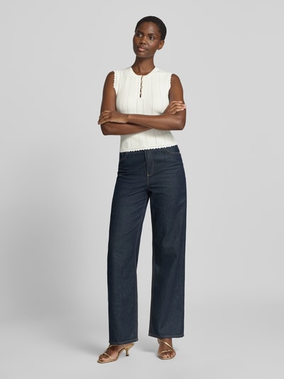 Frauenmodel trägt Stricktop in Pointelle-Strick mit kurzer Knopfleiste von GUESS in Weiß, körpernaher Schnitt, kombiniert mit dunkler Hose, Ganzkörperansicht von vorne.