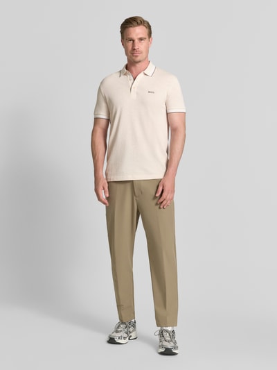 Męski model nosi kremową koszulkę polo Regular Fit Poloshirt aus reiner Baumwolle Modell 'PADDY' von BOSS Green, jasnobeżowe spodnie, szare buty sportowe, tło jednolite jasnoszare, Ganzkörperansicht von vorne.
