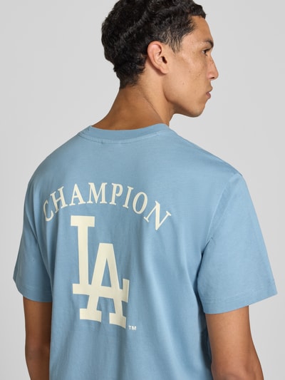 Männermodel trägt Relaxed Fit T-Shirt aus reiner Baumwolle von Champion in Hellblau mit Statement-Print und Label-Stitching, Ganzkörperansicht Rückansicht.
