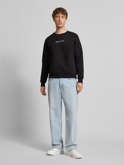 Męski model ma na sobie Sweatshirt mit Logo und Rundhalsausschnitt od Alpha Industries, czarny, bawełniany krój regular fit, logo, okrągły dekolt, długość tyłu 67 cm, długość rękawa 66 cm, pełen przód.