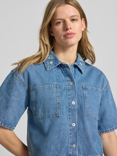 Frauenmodel trägt Boxy Fit Jeansbluse aus reiner Baumwolle Modell 'SKY' von Pieces, Denim-Optik, Stickerei, Brusttasche, Nahaufnahme Oberkörper.