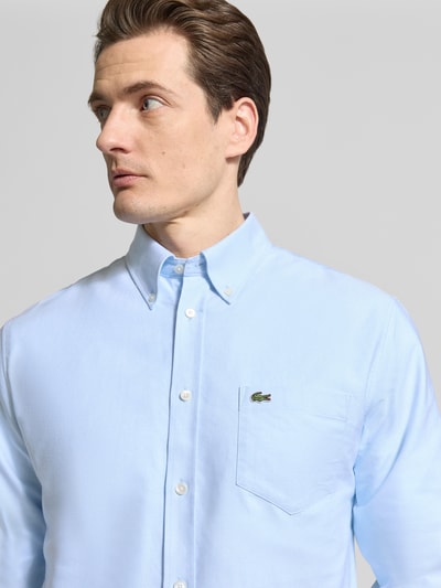 Męski model nosi Regular Fit Freizeithemd aus reiner Baumwolle od Lacoste, jasnoniebieski, logo na kieszeni, oxford, zbliżenie górnej części ciała.
