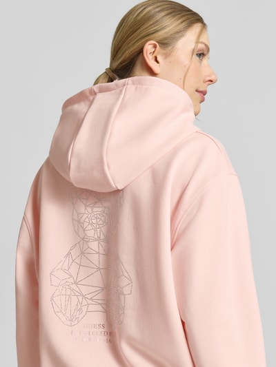 Vrouwenmodel draagt Oversized Hoodie met strassteendecoratie model 'Silvy' van Guess Activewear, lichtroze materiaal-mix met katoen, capuchon en kangoeroezak, elastische boorden, oversized fit, detailopname rugaanzicht.