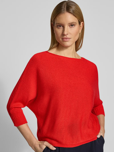 Kobieta modelka nosi sweter z dzianiny z dekoltem w łódkę i detalem z logo od More & More, prosty krój, kolor czerwony, długość tyłu 50 cm, długość rękawów 37 cm, zbliżenie na górną część ciała, widok z przodu.