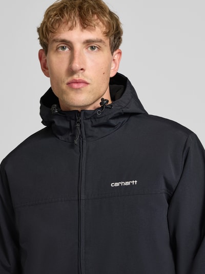 Männermodel trägt Funktionsjacke mit Logo-Stitching und Kapuze von Carhartt Work In Progress in Schwarz, glatte Textur, Nahaufnahme Oberkörper.