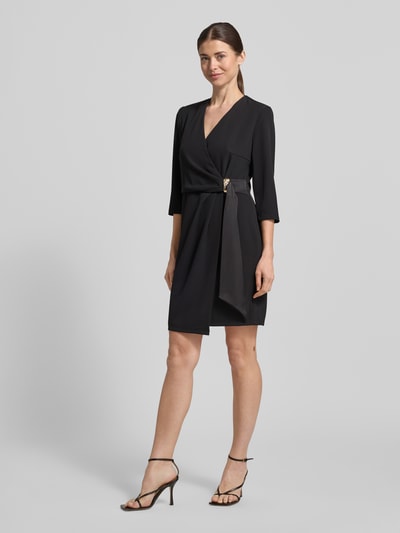 Modelka kobieca nosi Knielanges Cocktailkleid in Wickel-Optik od Rinascimento, mieszanka materiałów z dodatkiem streczu, prosty krój, długość do kolan, kopertowy styl, rękaw o dł. 3/4, długość tyłu w rozmiarze S: 90 cm, szerokość w rozmiarze S: 46 cm, długość rękawów w rozmiarze S: 44 cm, czarny kolor, gładka faktura, widok całej sylwetki z przodu