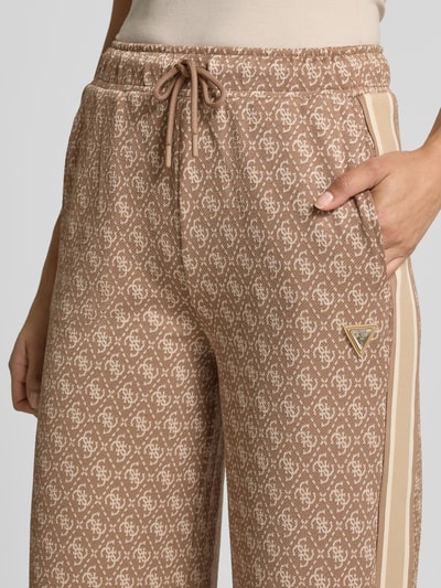 Frauenmodel trägt Sweatpants mit Logo-Muster im Allover-Look von GUESS in Beige und Creme, elastischer Bund, Nahaufnahme Oberkörper.