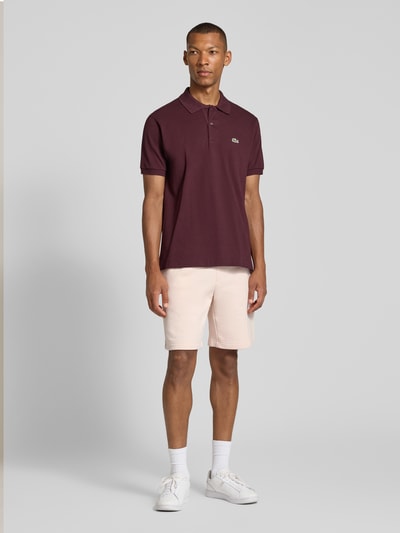 Mannenmodel draagt regular fit poloshirt uit puur katoen van Lacoste in donkerrood, effen, gecombineerd met lichte shorts, volledig vooraanzicht. Heren poloshirt van Lacoste, Regular Fit Poloshirt uit puur katoen.