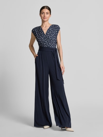 Vrouwenmodel draagt Regular Fit Jumpsuit met V-hals van Vera Mont, donkerblauw met witte stippen, elastische materiaalmix, mouwloos, getailleerde pasvorm, plooien, tailleband, ruglengte maat 36: 39 cm, borstwijdte: 44 cm, mouwlengte: 18 cm, totale lengte: 160 cm, binnenbeenlengte: 93 cm. Ganzlichaamsaanzicht van voren.