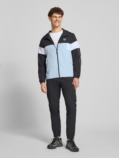 Męski model nosi Regular Fit Jacke mit Logo-Stitching Modell 'Caspio' od SERGIO TACCHINI, elastyczny materiał, kaptur, zamek, kieszenie, biało-czarno-niebieskie panele, długość tyłu 70 cm, długość rękawa 68 cm, widok całej sylwetki z przodu.