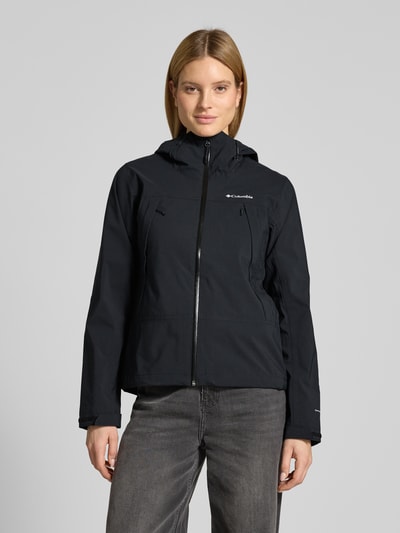 Frauenmodel trägt Regenjacke mit Omni-Tech™ ECO-Technologie von Columbia in Schwarz, Kapuze, Reißverschlusstaschen, verstellbare Ärmelriegel, leicht taillierte Passform, atmungsaktiv, wasserdicht, Futter aus Mesh und PU-Beschichtung, Ganzkörperansicht von vorne.