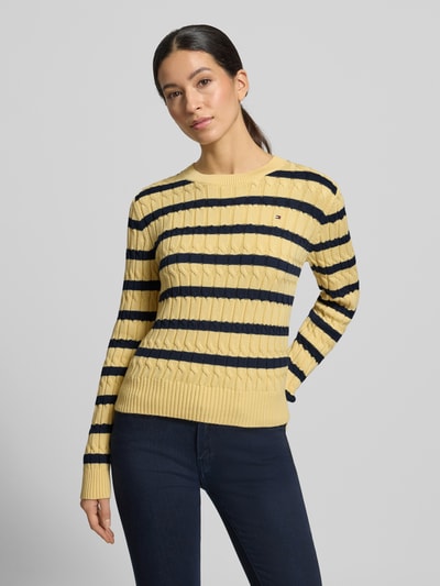 Frauenmodel trägt Slim Fit Strickpullover aus reiner Baumwolle von Tommy Hilfiger in Gelb mit schwarzen Streifen, Logo-Detail, Crew Neck, gerippte Abschlüsse, Zwei-Ton-Optik, Ganzkörperansicht von vorne.