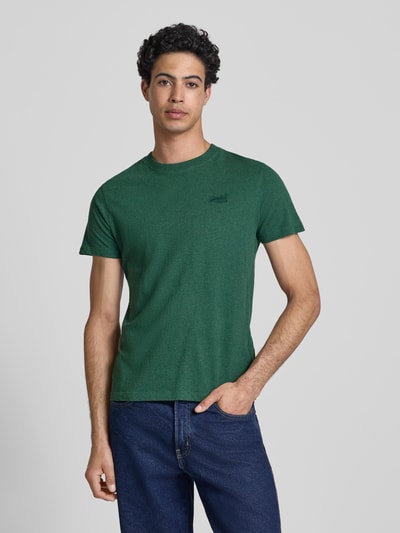 Männermodel trägt Regular Fit T-Shirt mit Logo-Stitching von Superdry in Dunkelgrün, reine Baumwolle, Rundhalsausschnitt, Jeans, Ganzkörperansicht von vorne.