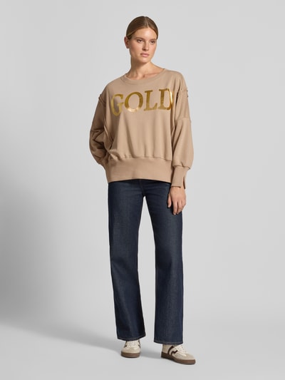 Frauenmodel trägt Oversized Sweatshirt aus reiner Baumwolle mit Statement-Print von miss goodlife in Beige mit goldfarbenem Schriftzug, kombiniert zu Jeans und Sneakers, Rundhalsausschnitt und elastische Abschlüsse, Ganzkörperansicht von vorne.