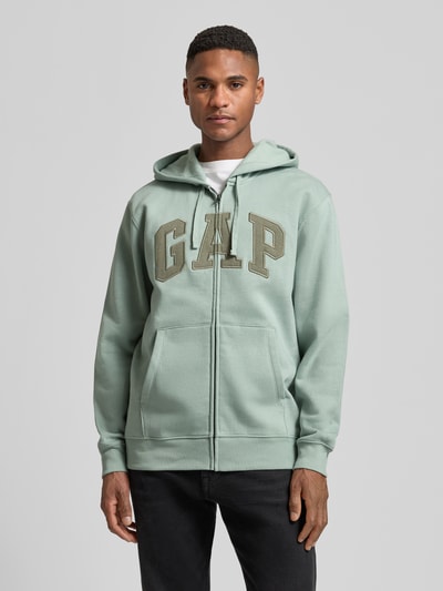 Männermodel trägt Sweatjacke mit Logo und Känguru-Tasche von GAP in Salbeigrün, Baumwoll-Mix, Regular Fit, Ganzkörperansicht von vorne.