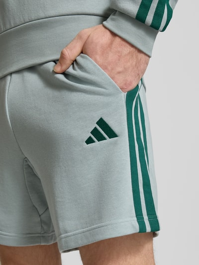 Männermodel trägt Regular Fit Shorts mit Viskose-Anteil von adidas Sportswear, hellgrün mit dunkelgrünen Streifen, Nahaufnahme Oberkörper. Herren Shorts, Baumwoll-Mix, elastischer Bund mit Tunnelzug, Logo-Stitching, Bundweite Größe M 40 cm.