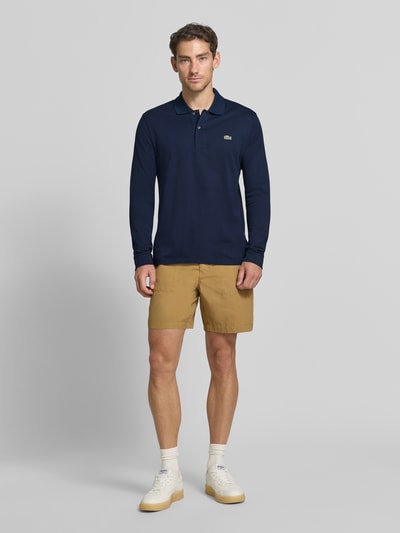 Männermodel trägt Regular Fit Poloshirt aus reiner Baumwolle von Lacoste in Dunkelblau, kombiniert mit beigefarbenen Shorts, Ganzkörperansicht von vorne, Herren Poloshirt von Lacoste.