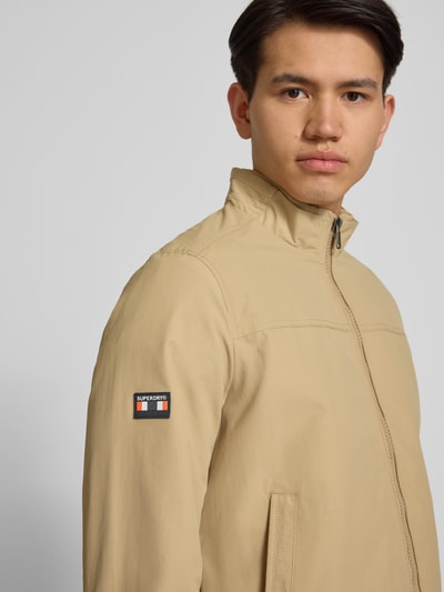 Męski model nosi Jacke mit Stehkragen und Eingrifftaschen od Superdry, beż, wpuszczane kieszenie, krój regular fit, stójka, zamek błyskawiczny, długość tyłu w rozmiarze M: 70 cm, szerokość ramion w rozmiarze M: 44 cm, szerokość klatki piersiowej w rozmiarze M: 41 cm, długość rękawów w rozmiarze M: 68 cm, zbliżenie na górną część ciała z przodu