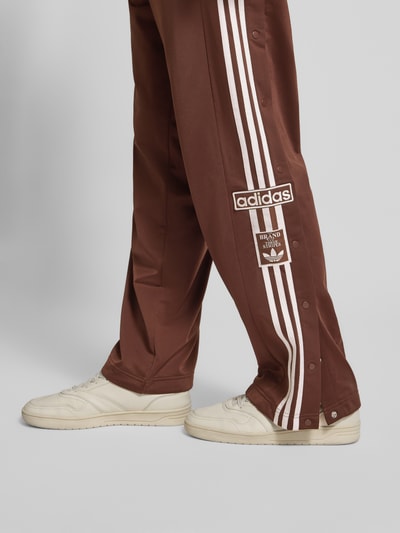 Männermodel trägt Wide Fit Jogginghose mit Logo-Stitching Modell 'Adibreak' von adidas Originals, Knopfleisten, Logo-Patch, Nahaufnahme Bein.