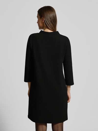 Frauenmodel trägt Minikleid mit 3/4-Ärmel von More & More, schwarzer Material-Mix mit Baumwoll-Anteil, Struktur-Oberfläche, Ganzkörperansicht Rückansicht.