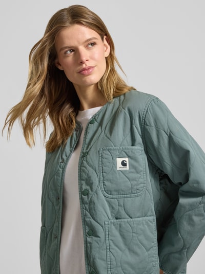 Vrouwenmodel draagt Steppjacke mit Label-Detail Modell 'Kylan' van Carhartt Work In Progress in relaxed fit, katoenen stof, ruittextuur, opgestikte zakken, ronde hals, knoopsluiting, labeldetail, voorkant bovenlichaam close-up.