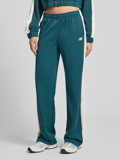 Frauenmodel trägt Sweatpants mit elastischem Bund und Label-Stitching von New Balance in Petrol mit weißen seitlichen Streifen, Relaxed-Fit, Eingrifftaschen, Innenbeinlänge bei Größe S 80 cm, Nahaufnahme Unterkörper, Ganzkörperansicht von vorne.