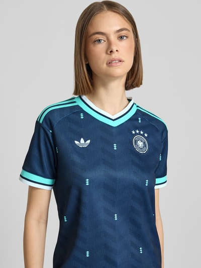 Modelka nosi DFB Auswärtstrikot 2026 od adidas Sportswear, wielokolorowe, krój regular fit, logo DFB, dekolt w serek, krótki rękaw, detalowe przeszycia z logo, tekstura o falistym wzorze; zbliżenie na górną część ciała z przodu.