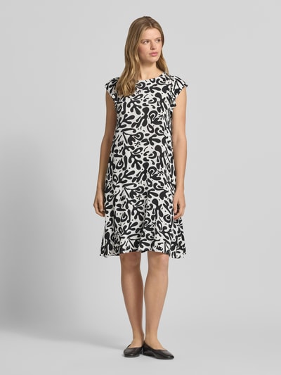 Frauenmodel trägt Regular Fit Sommerkleid aus reiner Viskose Modell 'BARRI' von Pieces mit Allover-Muster in Schwarz-Weiß, knielang, Rundhalsausschnitt, kurzarm mit Schmetterlingsärmeln, Crêpe-Textur, Ganzkörperansicht von vorne.