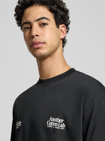 Männermodel trägt T-Shirt mit Label-Print von Another Cotton Lab in Schwarz, Reine Baumwolle, Lockerer-Schnitt, Nahaufnahme Oberkörper.